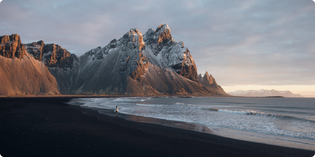 A dramatic landscape photograph of a black sand beach with towering jagged mountains, some dusted with snow, under a cloudy sky at sunrise or sunset.